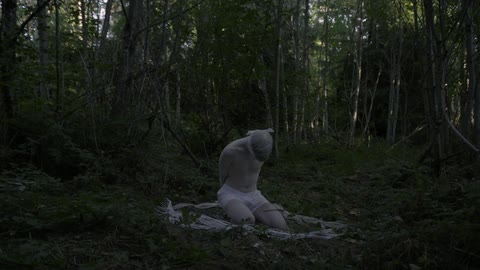 Pale blindfolded man in white boxers kneels on the forest ground surrounded by white straw