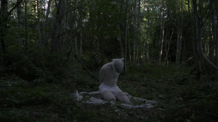 Pale blindfolded man in white boxers kneels on the forest ground surrounded by white straw