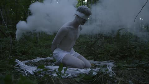 Smoke billows behind a blindfolded man in the woods