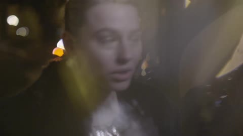 A woman wearing a shiny gold dress and fur coat walking and talking then stopping to put her hand to her forehead