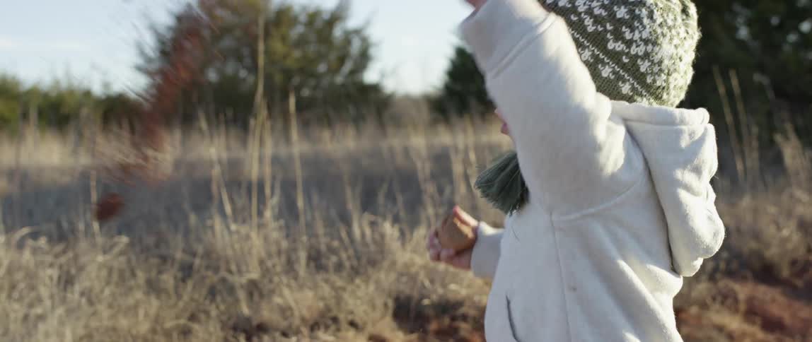 A toddler standing in a field, holding a rock in one hand and throwing something with the other