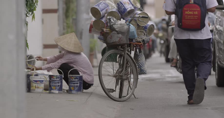 A person squatting and pouring something from one paint can into another next to a bicycle with many paint cans tied to the frame