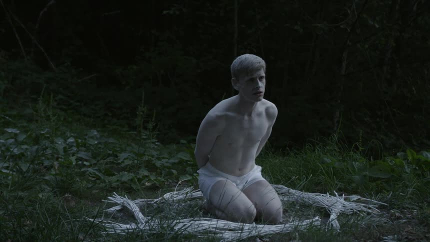 A very pale man, dressed only in white boxers and dusted with white powder, kneeling in the middle of a circle of logs in a wooded area