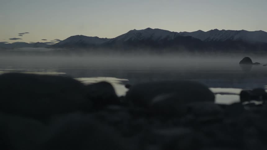 Fog hovers over a still lake that is surrounded by black rock