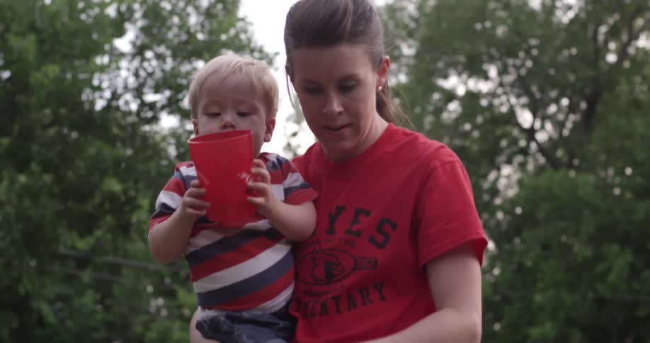 A woman walks while holding a toddler who is holding a large red cup