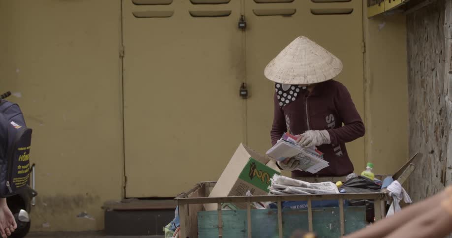 A person rummages through trash while pedestrians wearing face masks pass by