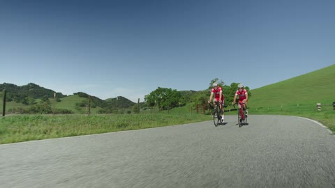 Two men riding ten speeds down a winding rural road with hills in the background