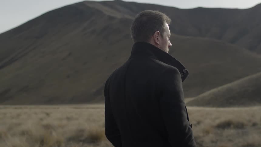 A man in a pee coat stands staring across a plain towards some hills