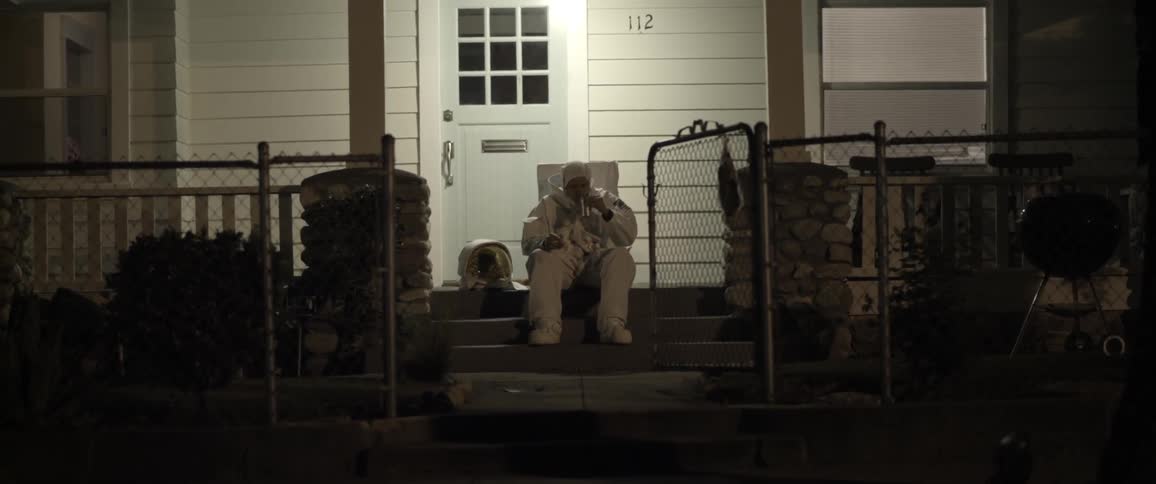 A person wearing an astronaut suit, sits on front porch drinking
