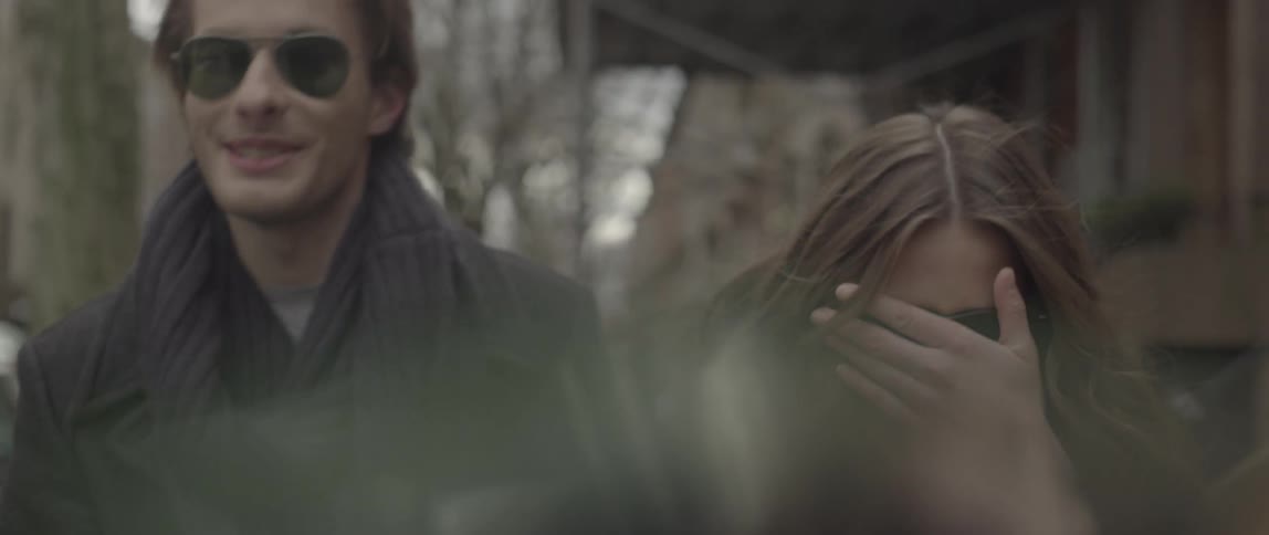A man and woman are walking down the street, but she has her hand over her eyes
