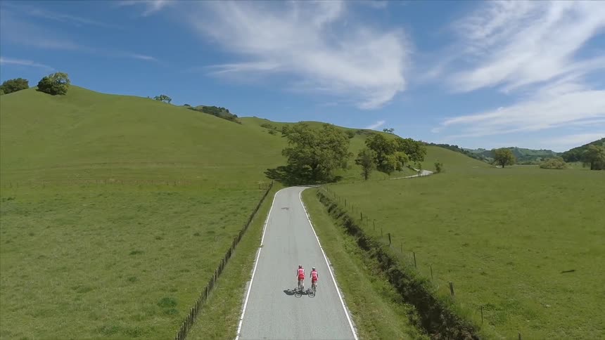 Two people ride bicycles down a road in a hilly, verdant country setting