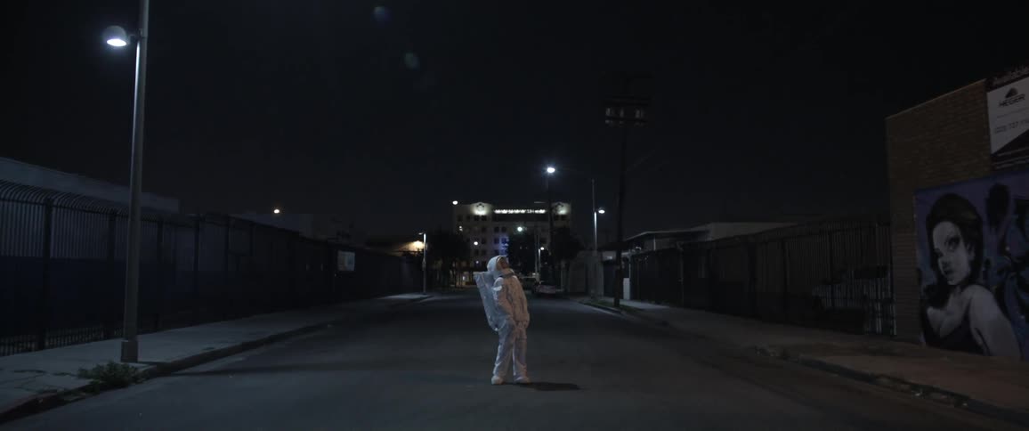 A person in an astronaut suit stands in the middle of an empty street at night and looks toward the sky