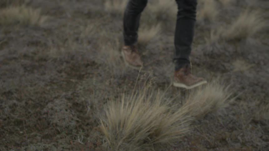 The legs and feet of a man walking over tufts of grass in a natural setting