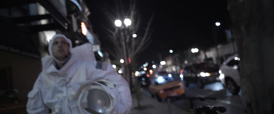 Man in astronaut suit walks down a city sidewalk at night