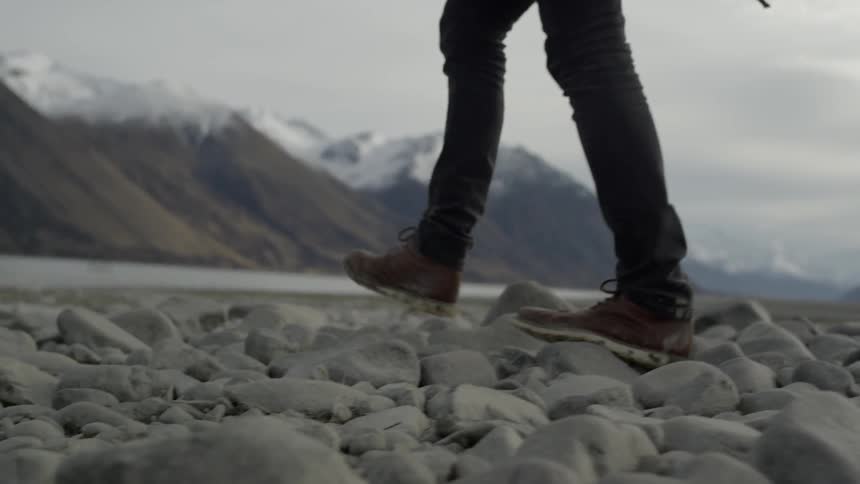A person walks across rocks near water and a mountain