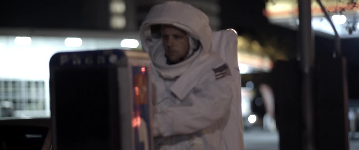 A man in an astronaut costume uses a phone booth at a gas station