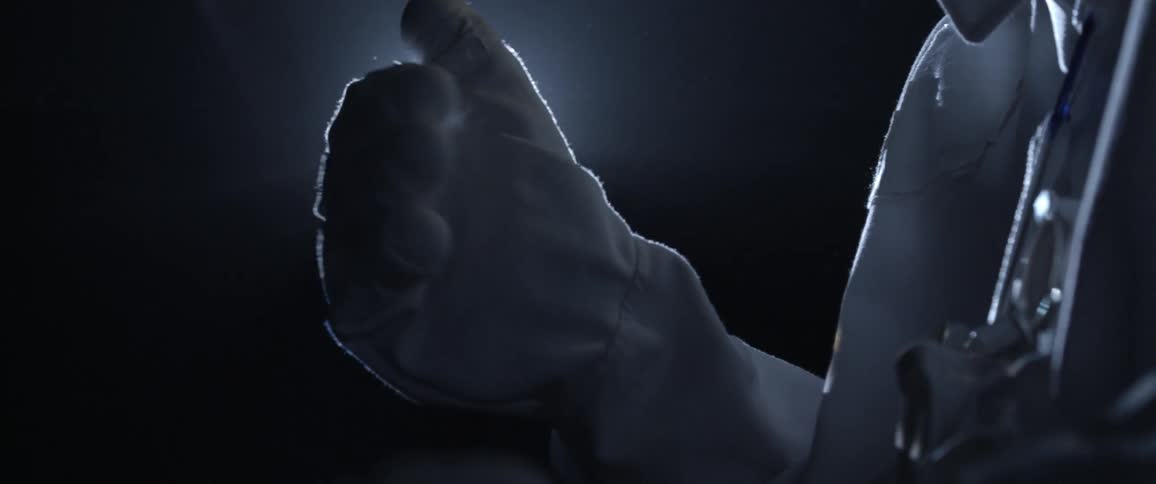 A man dressed as an astronaut examines his hands in gloves in front of a bright light