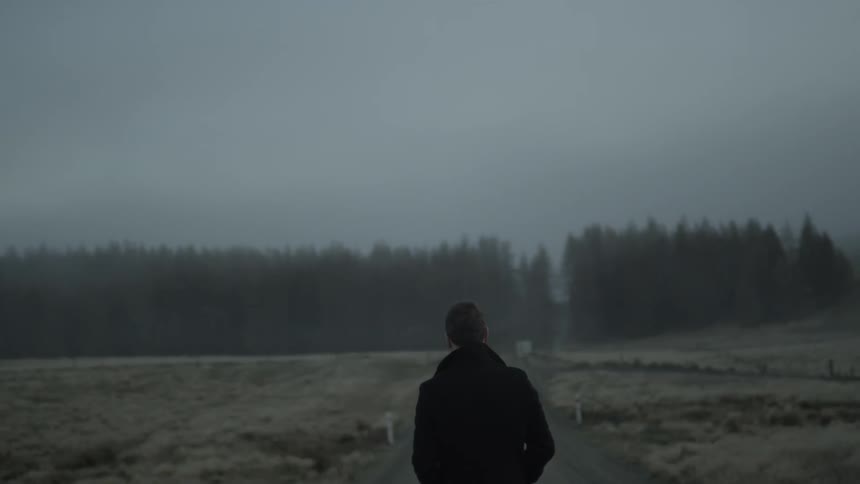 A white man in a dark coat walks down the center of a dirt road in a frost covered landscape
