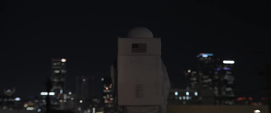 A person in a white astronaut costume with an american flag on the back stands and looks at a city at night