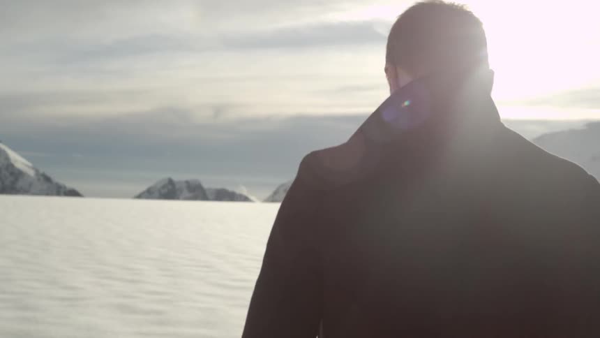 A young man in a dark coat walks in an icy landscape towards some mountains