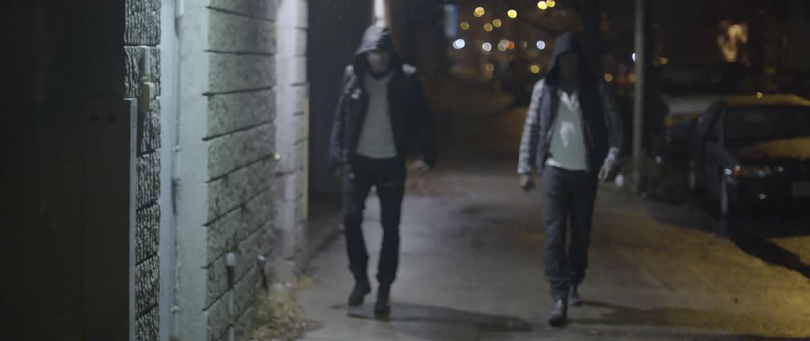 A man walks down a street at night while being followed by two men in masks