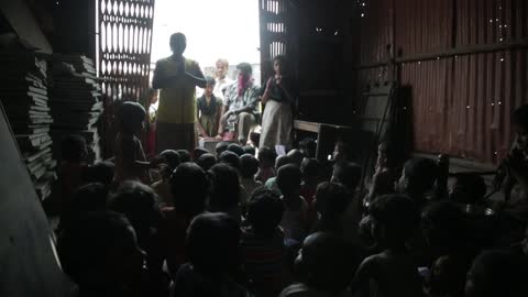 A group of children in a dark room listen to a teacher speaking