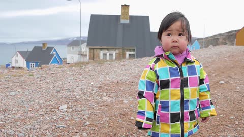 A little girl in a brightly colored jacket stands on a slope with homes behind her