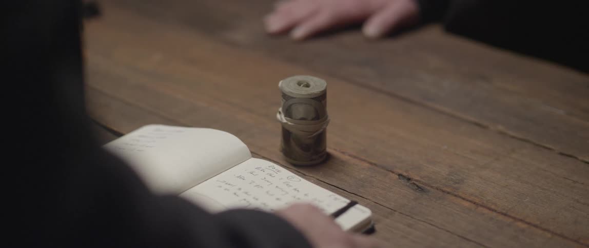 A roll of cash is placed down on a wooden table in front of a person with an open ledger after a pause the person picks up the roll of money