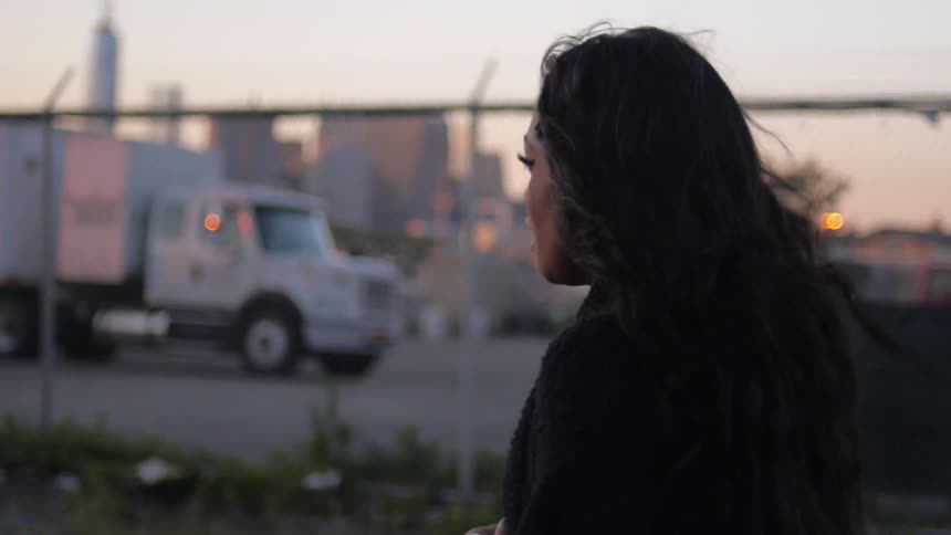 Woman walking down a city street next to a fence with a semi in a parking lot behind it