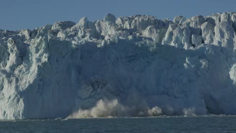 Snow and ice fall from the side of a mountain and collapse into a large body of water