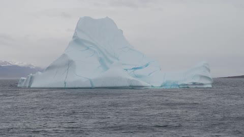 An iceberg in the sea with snow-capped mountains in the background