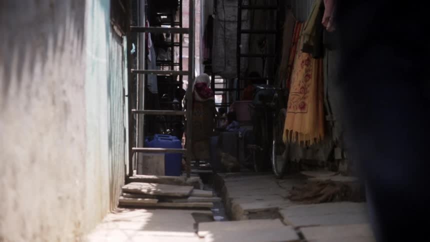 A girl walks toward her mother and brother in an alley as they feed chickens and hang laundry