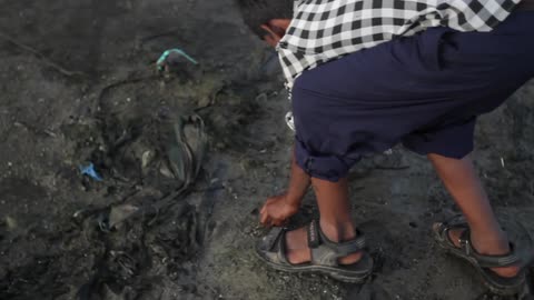 A young boy rolls up his pant legs picks up a rock and throws it across a dirt soil terrain