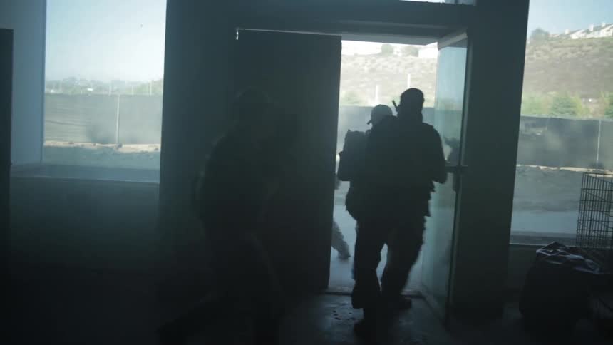 A military unit with weapons drawn and aimed escorts civilians out of a building