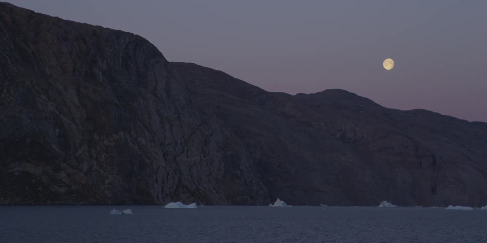 A rocky mountain sitting on an arctic body of water during nightfall