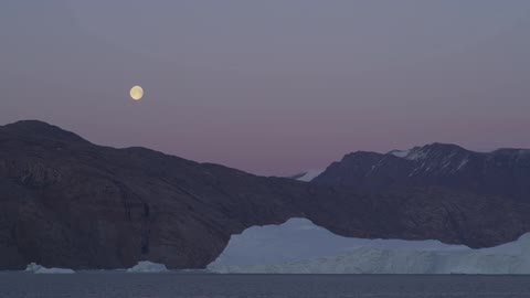 The moon is shown at dusk above a snow covered mountain range