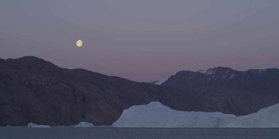 The moon is shown at dusk above a snow covered mountain range