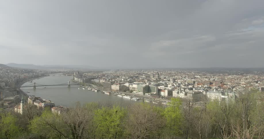 An older towns skyline with a bridge that runs over the river surrounded by budding green trees under a stormy sky