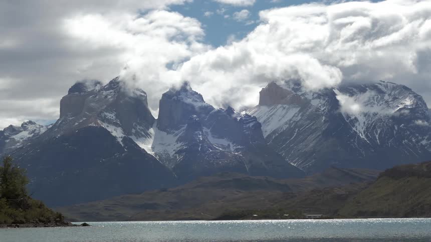 Clouds blowing over the tops of snow-covered mountain peaks behind a body of water
