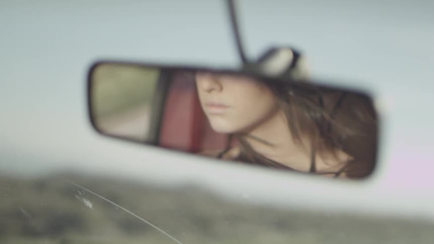 A woman drives down the road as her reflection is shown in the rearview mirror