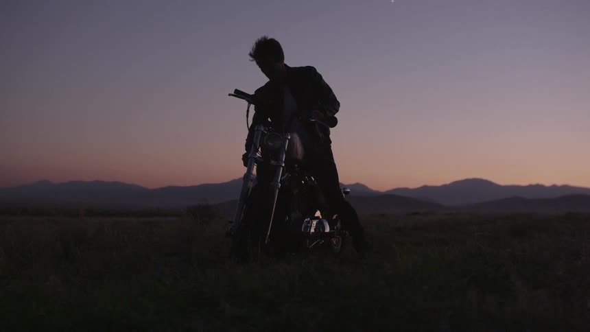 A man sits on a motorcycle in a field at dusk