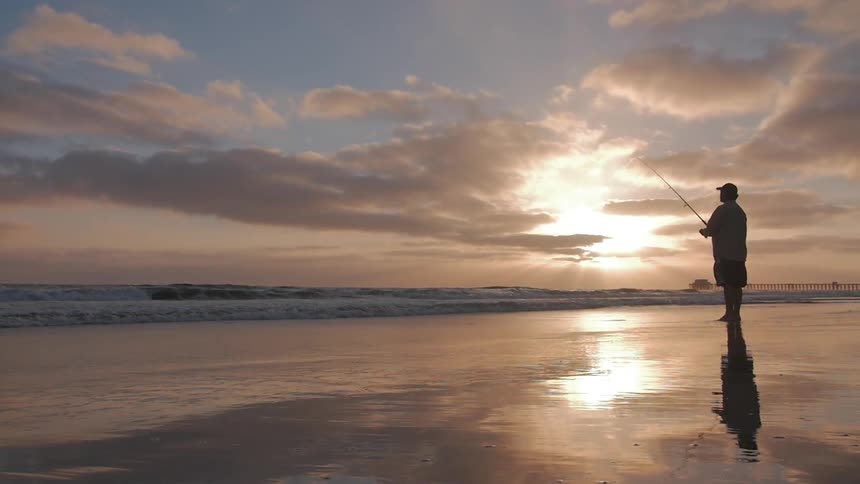A man fishes off the coast of a beach at sunset