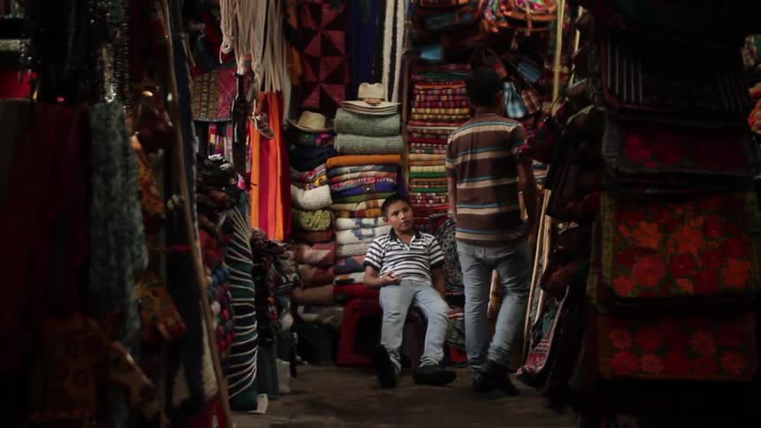 A boy sits in a shop filled with colorful fabrics as another boy walks past him