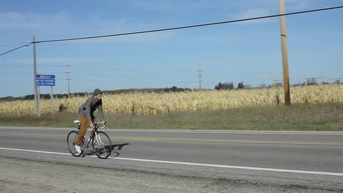 A man bicycles down a road passing a corn field