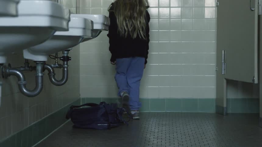 A little girl throws her backpack down in a public restroom then sits on the floor in the corner