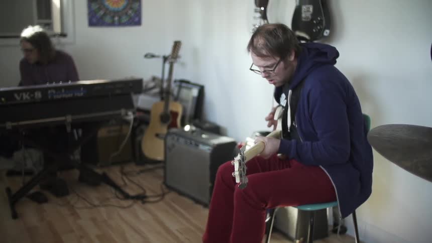 One man plays guitar, another plays keyboard sitting in a room