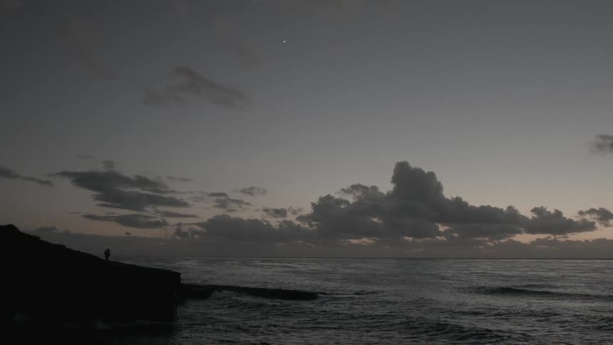 A view of waves crashing against a cliff on a cloudy evening while someone stands at the end of the cliff