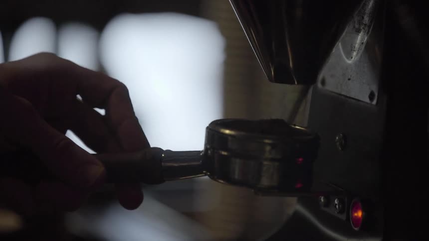 A hand holds an espresso portafilter under coffee grinder