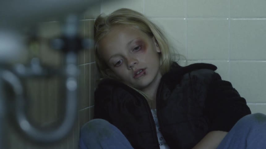 A young, blonde girl with a black eye and cut lip sitting in the corner on a bathroom floor
