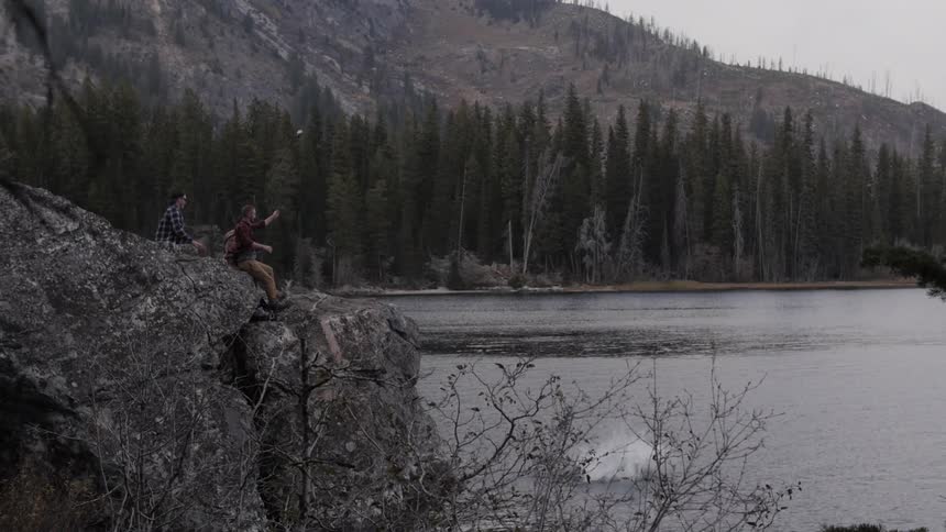 People sitting on a cliff as a man jumps off the cliff and into a body of water that is surrounded by trees and has mountains in the background